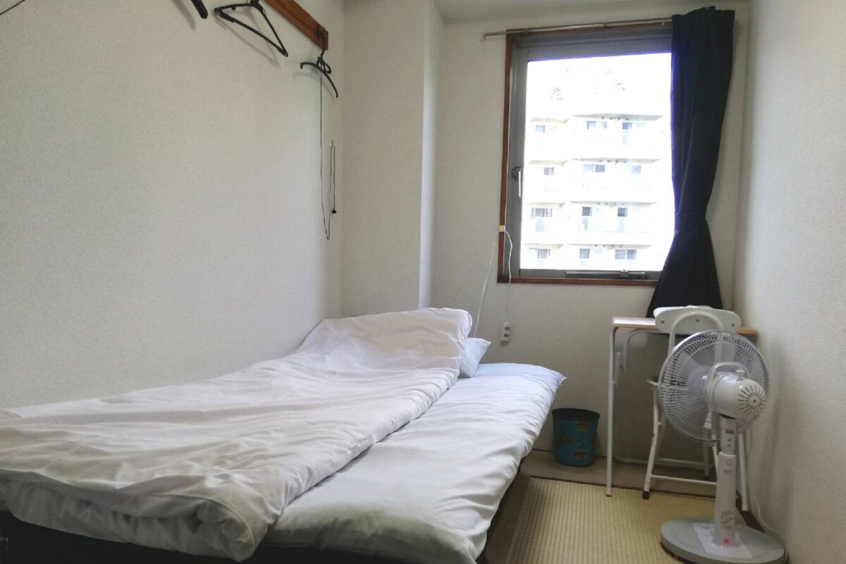 A small, single-bed room with a window, a standing fan, and a waste bin.