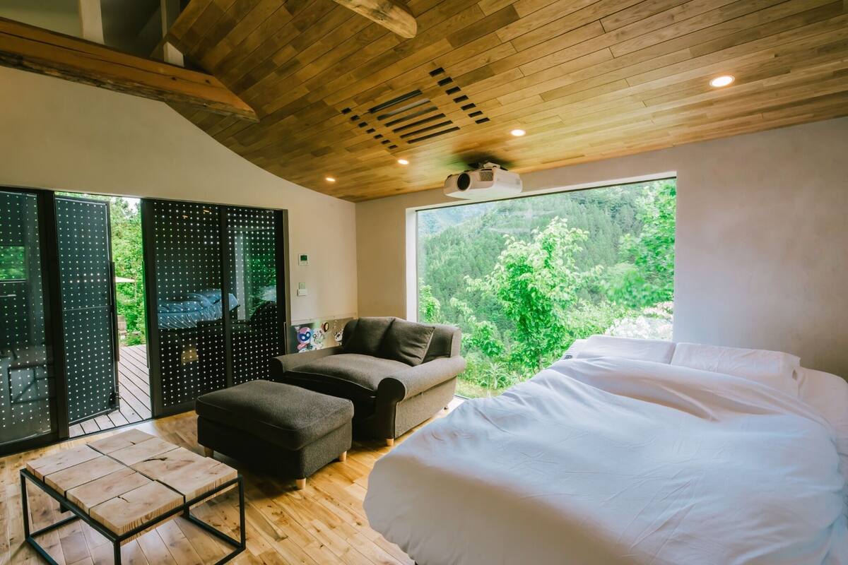 A bedroom with a large bed, a sofa, a small table, and a view of greenery through a window.