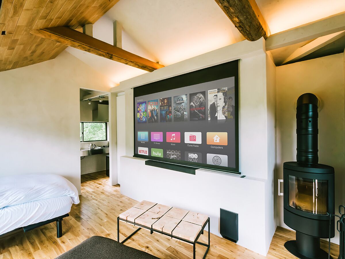 A cozy room with a bed, a small table, a wall-mounted TV displaying various apps, and a wood-burning stove.