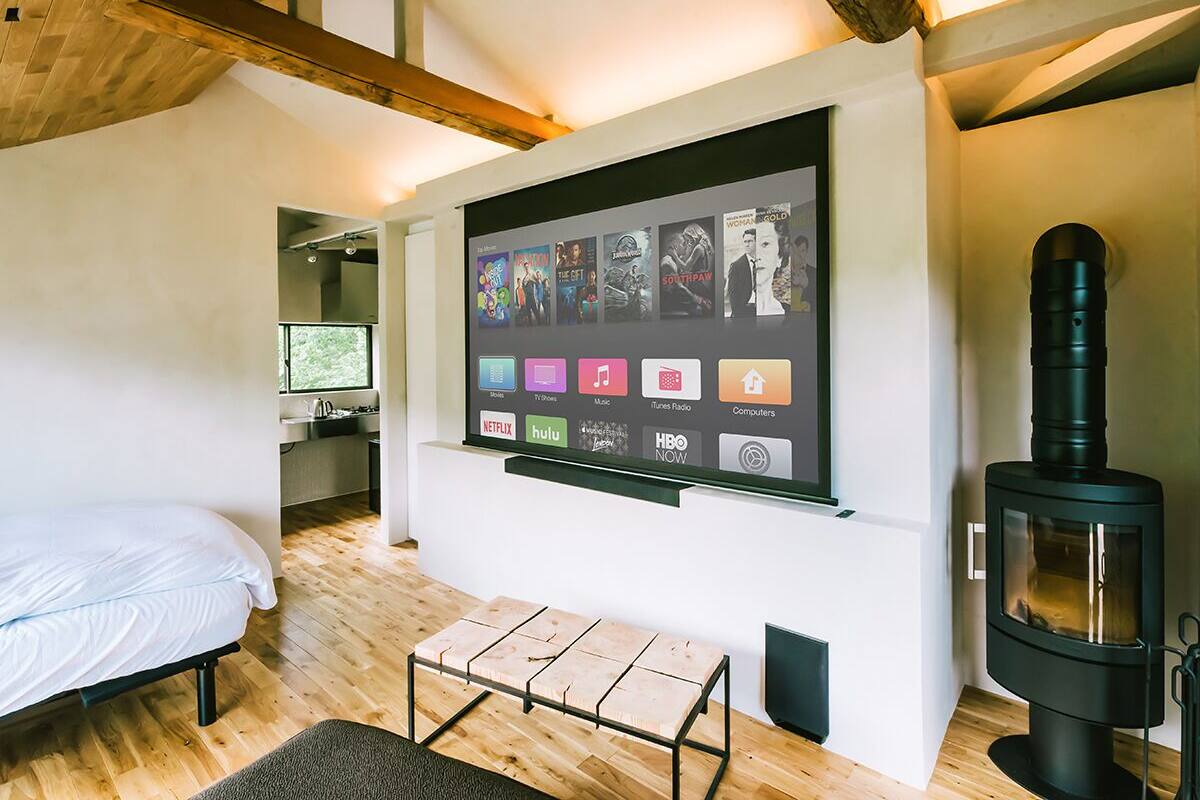 A cozy room with a bed, a small table, a wall-mounted TV displaying various apps, and a wood-burning stove.