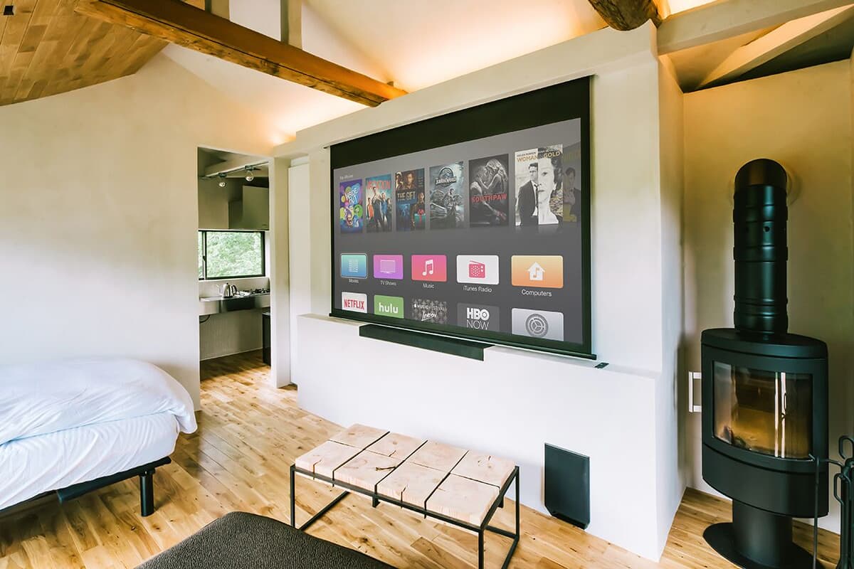 A cozy room with a bed, a small table, a wall-mounted TV displaying various apps, and a wood-burning stove.