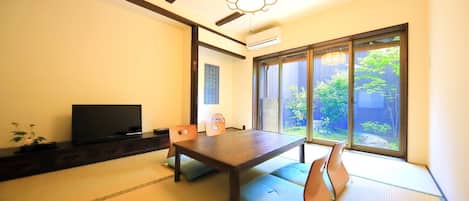 Una habitación tradicional japonesa con suelo de tatami, una mesa baja, un televisor y una puerta corredera de vidrio que da a un jardín.