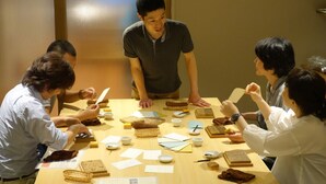 A group of people seated around a table with various items, including papers, a laptop, and small containers.