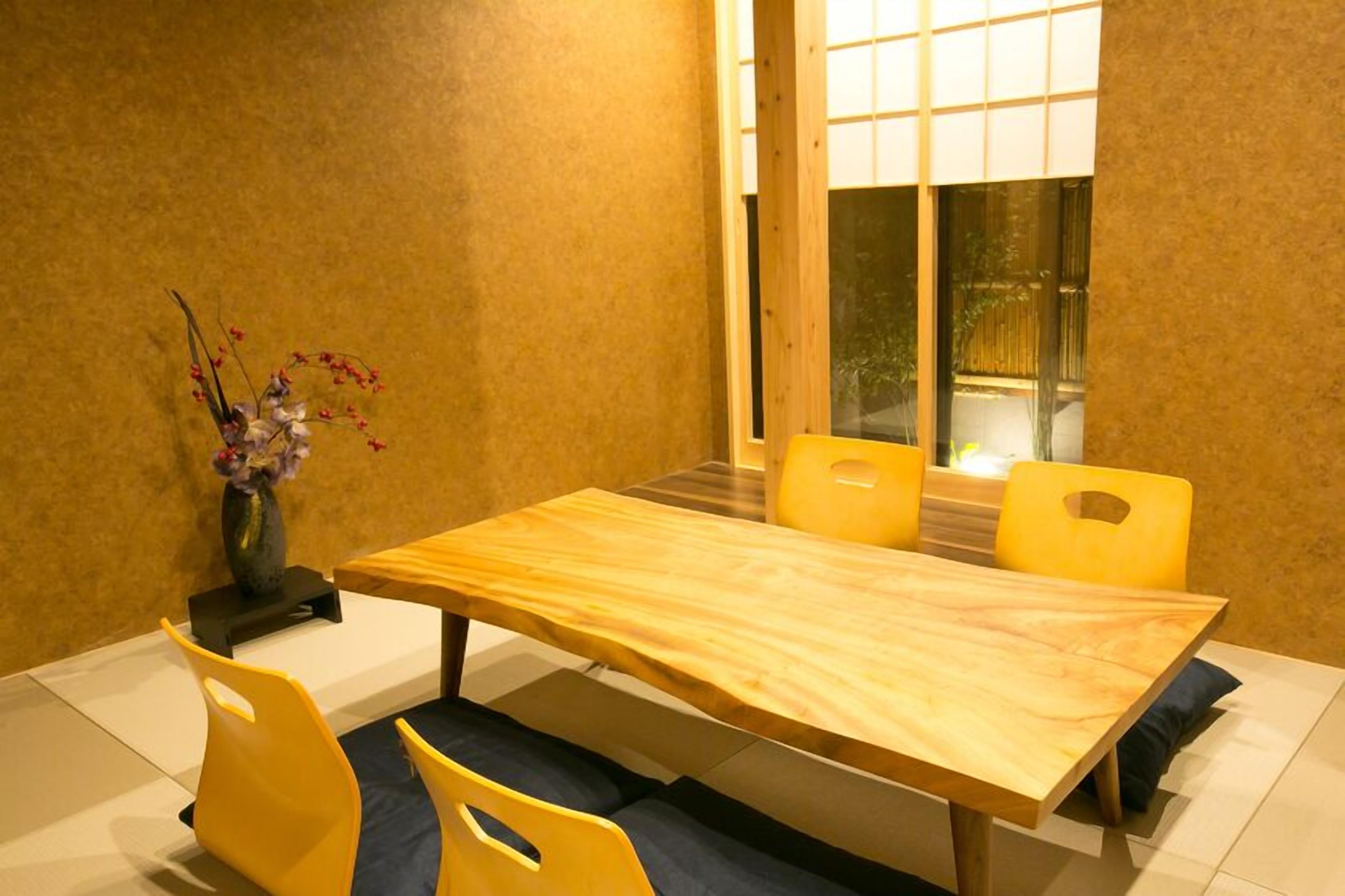 Een eethoek met een houten tafel en gele stoelen, een vaas met bloemen, en een raam met uitzicht op groen.