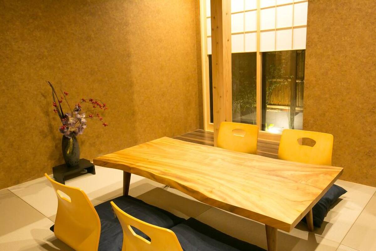 A dining area with a wooden table and yellow chairs, a vase with flowers, and a window with a view of greenery.
