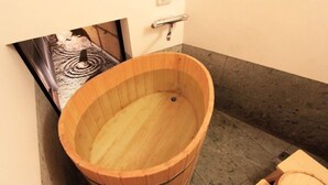 Eine hölzerne Badewanne in einer Ecke des Raumes mit Fliesenboden und einem kleinen Holzschemel.