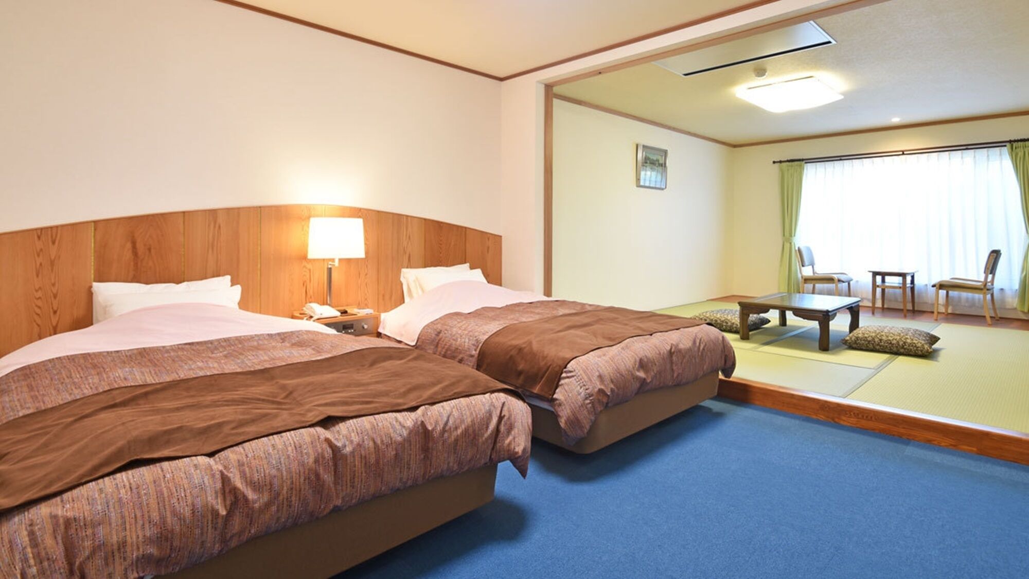 Una habitación de hotel con dos camas, una mesita de noche con lámpara, una pequeña mesa y un espacio de descanso con sillas y sofá.