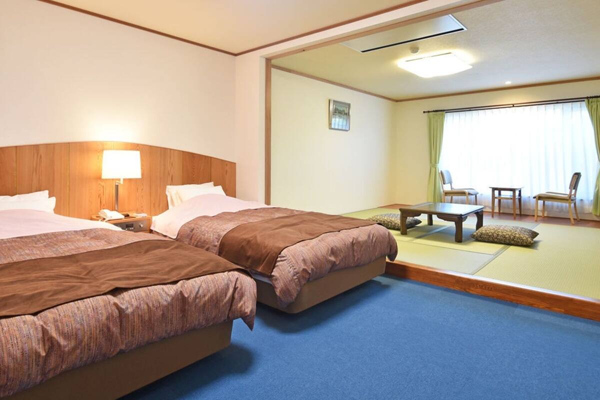 Una habitación de hotel con dos camas, una mesita de noche con lámpara, una pequeña mesa y un espacio de descanso con sillas y sofá.