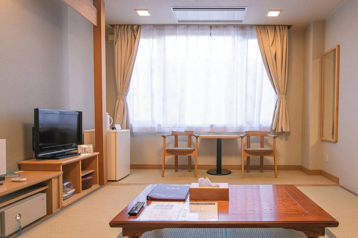 Habitación de hotel con televisor, una pequeña mesa con sillas, ventana con cortinas y un espejo en la pared.
