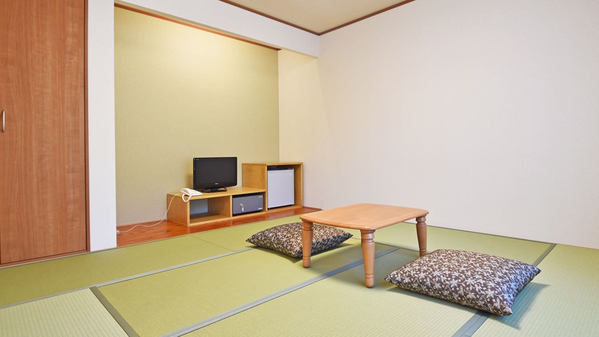 Ein Raum mit Tatami-Matten, einem niedrigen Holztisch, Kissen, einem Fernseher und einem kleinen Schrank.