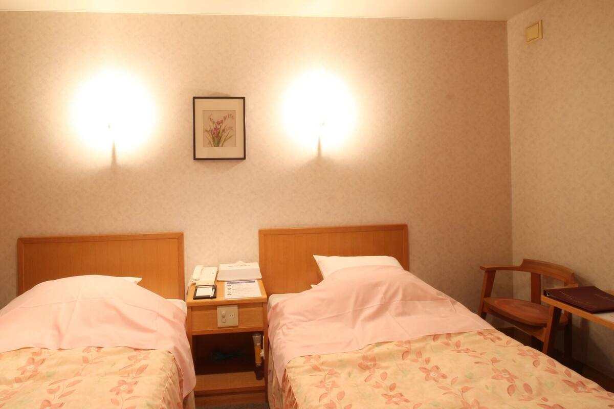 Habitación de hotel con dos camas individuales, una mesita de noche con teléfono y lámpara, un cuadro enmarcado en la pared y una silla de madera.