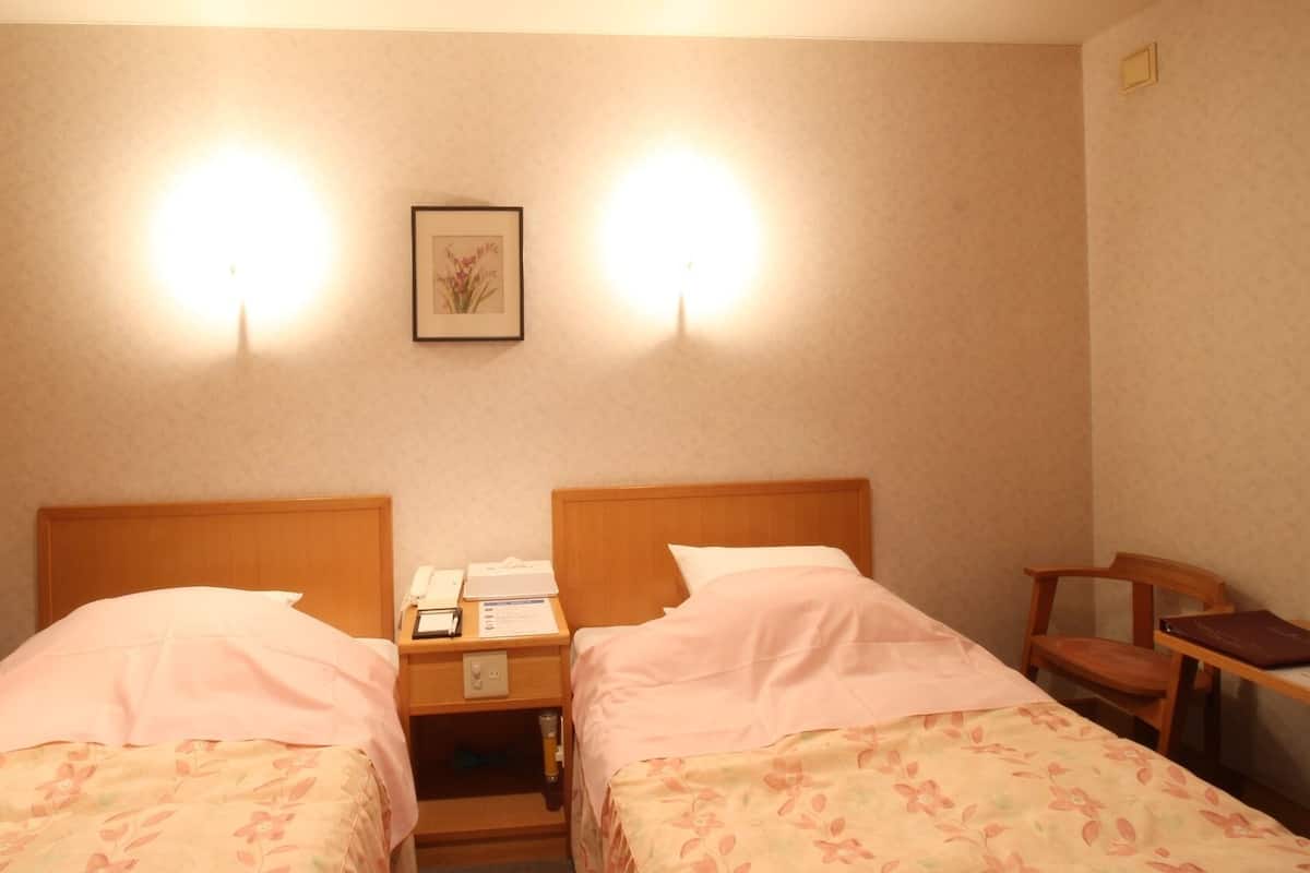 Een hotelkamer met twee eenpersoonsbedden, een nachtkastje met een telefoon en een lamp, een ingelijst schilderij aan de muur, en een houten stoel.