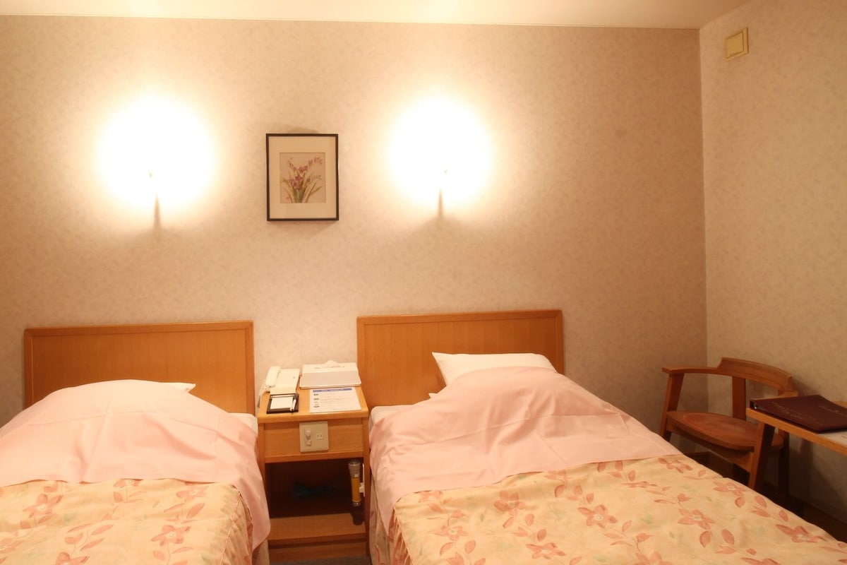 Habitación de hotel con dos camas, una mesita de noche con un teléfono y una lámpara, un cuadro en la pared y una silla.