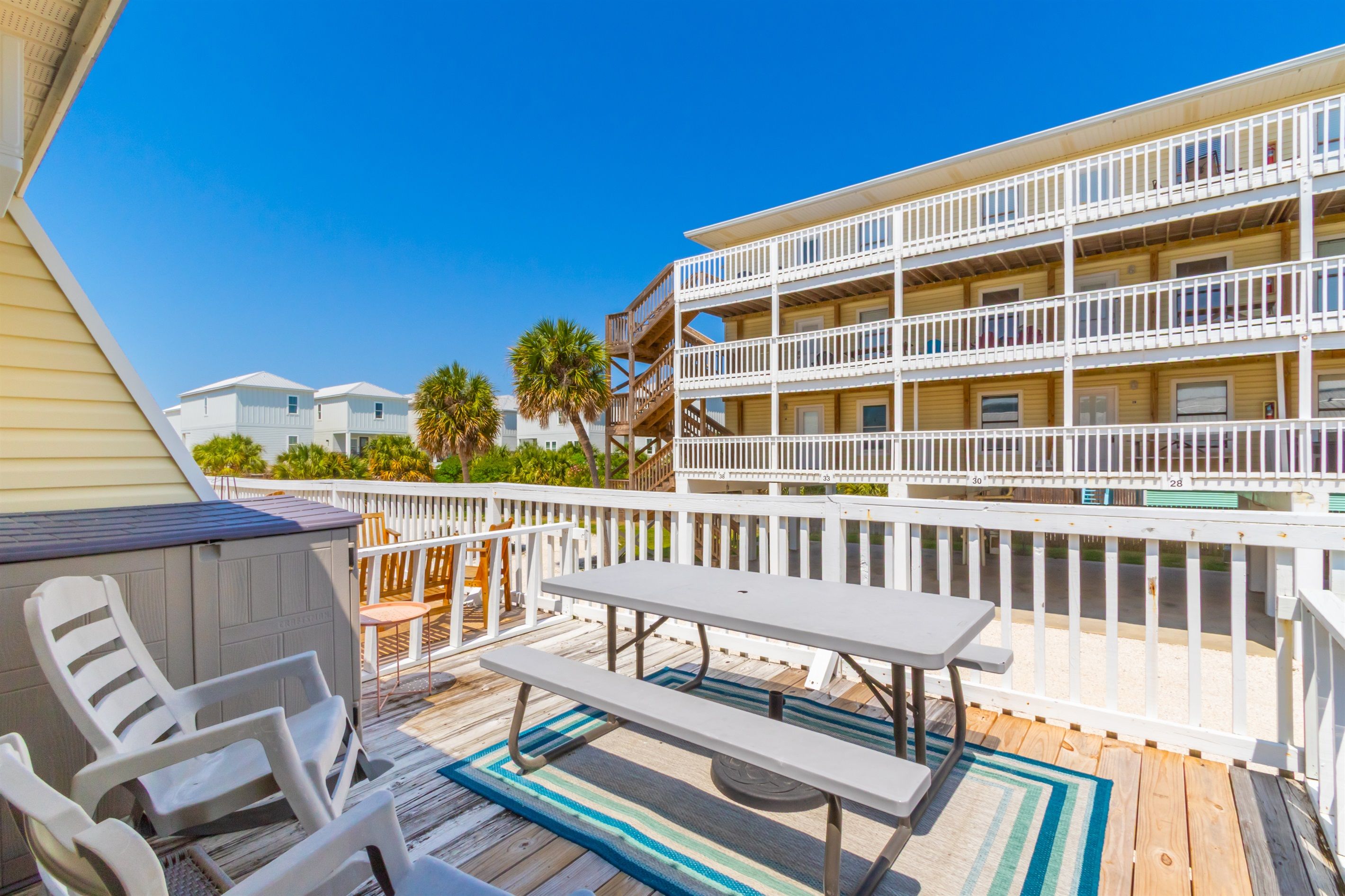 A deck with white chairs and a picnic table, overlooking a multi-story building with balconies.