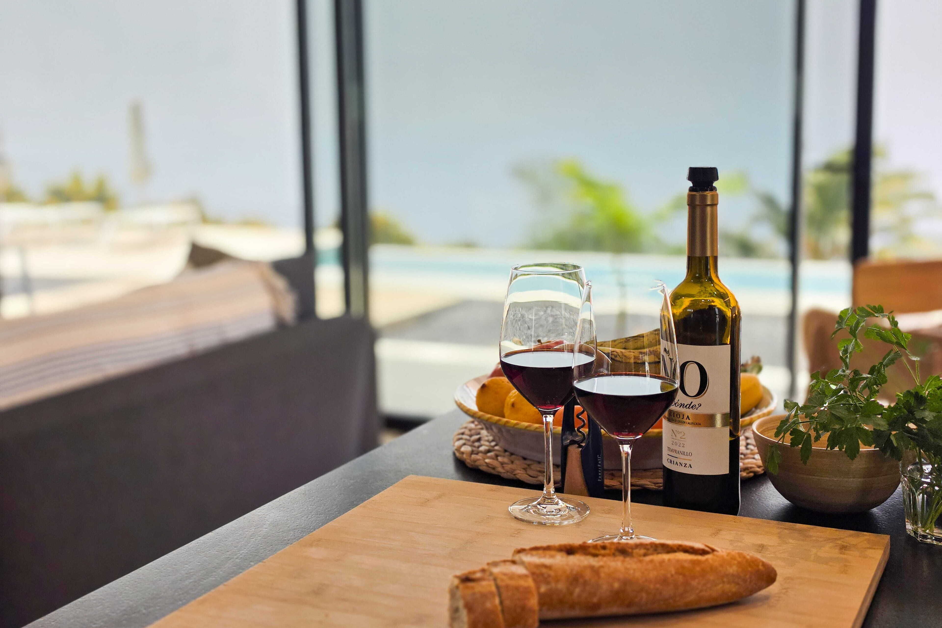 A table with a bottle of wine, glasses, bread, and a bowl of fruit by a window overlooking a pool.