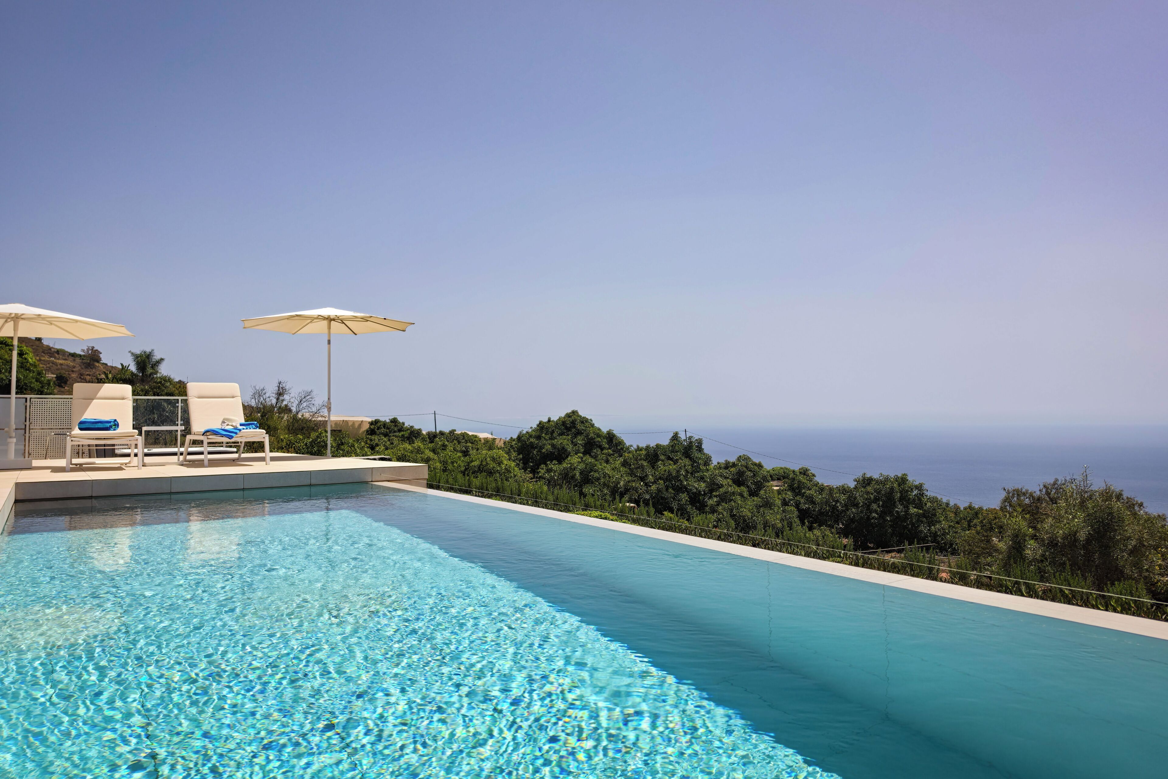 Une piscine d’un bleu éclatant, bordée de chaises longues et d’ombrelles, entourée de verdure et donnant sur une étendue d’eau.