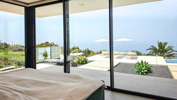 Een moderne slaapkamer met een groot bed, een schuifglasdeur naar een balkon met buitenmeubilair en een zwembad.