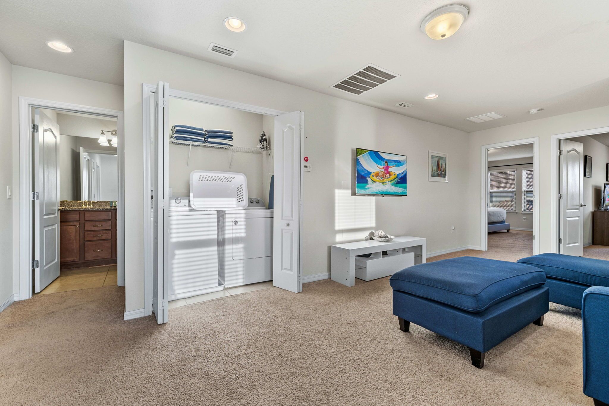 Una habitación amplia con un ventanal grande, un escritorio blanco, un sofá azul y una puerta que da a otra habitación.