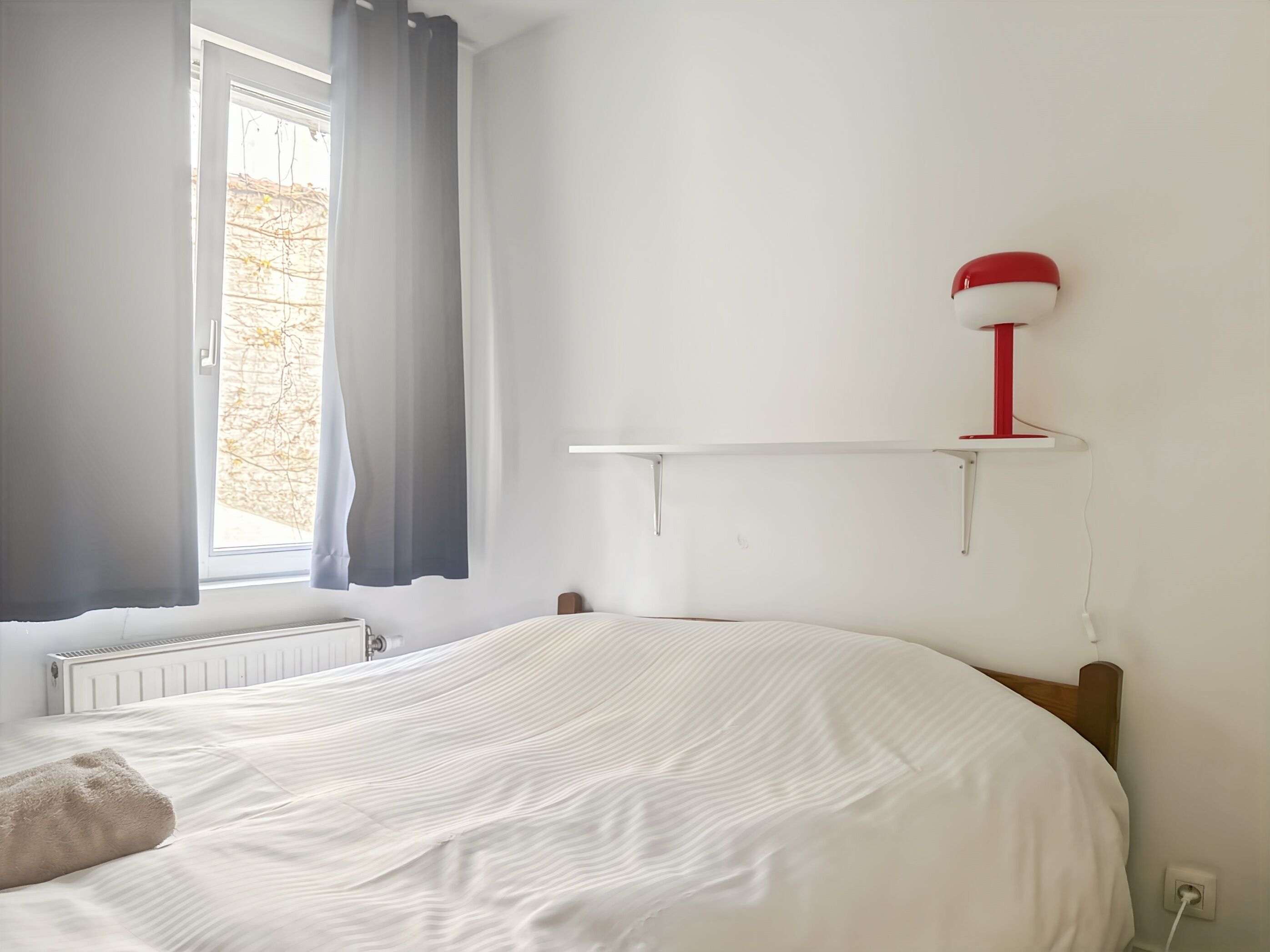 Quarto com cama, radiador, janela com cortinas e abajur vermelho e branco sobre uma prateleira branca.