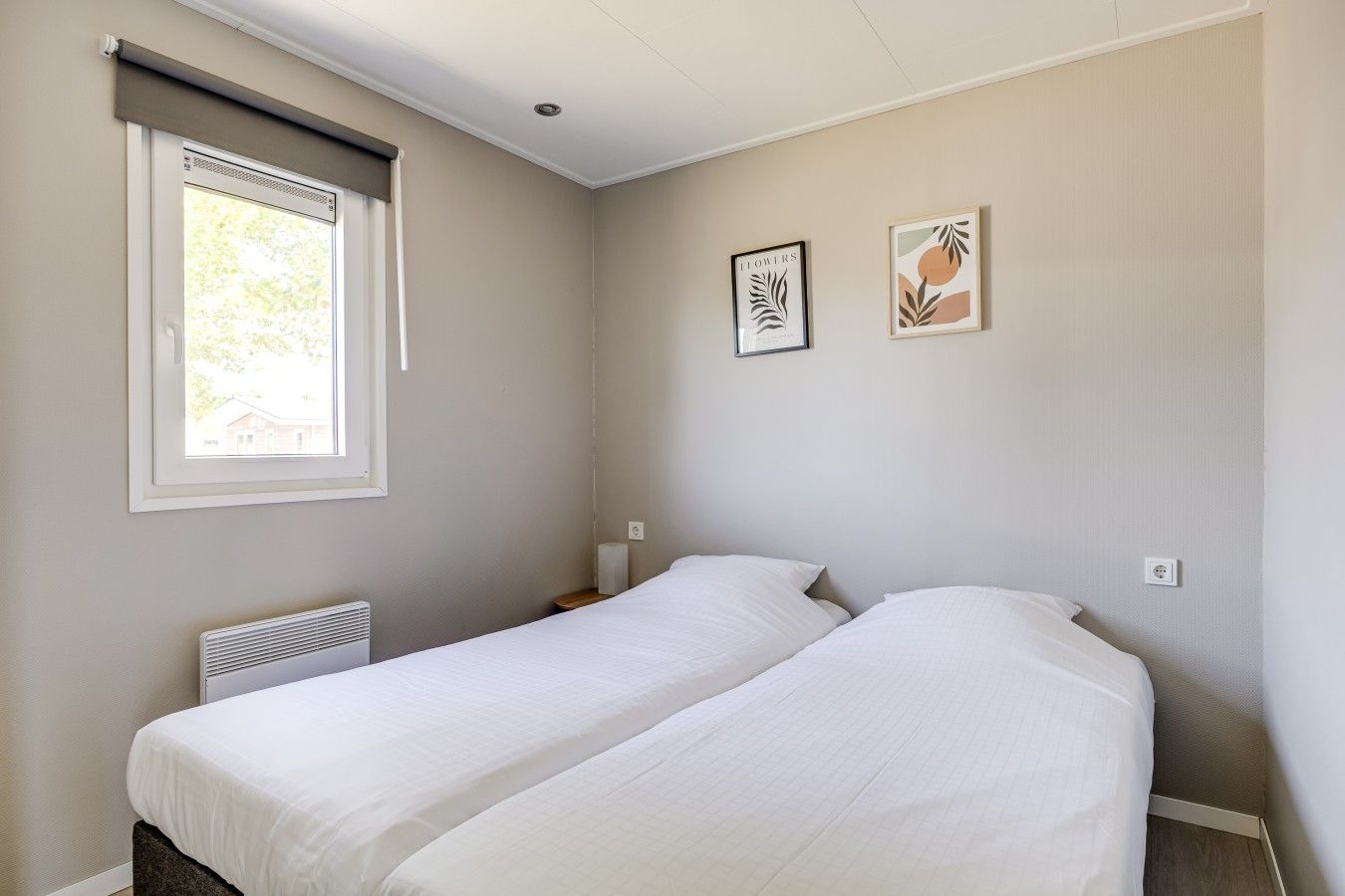 A room with two single beds, a window with a blind, a radiator, and two framed pictures on the wall.