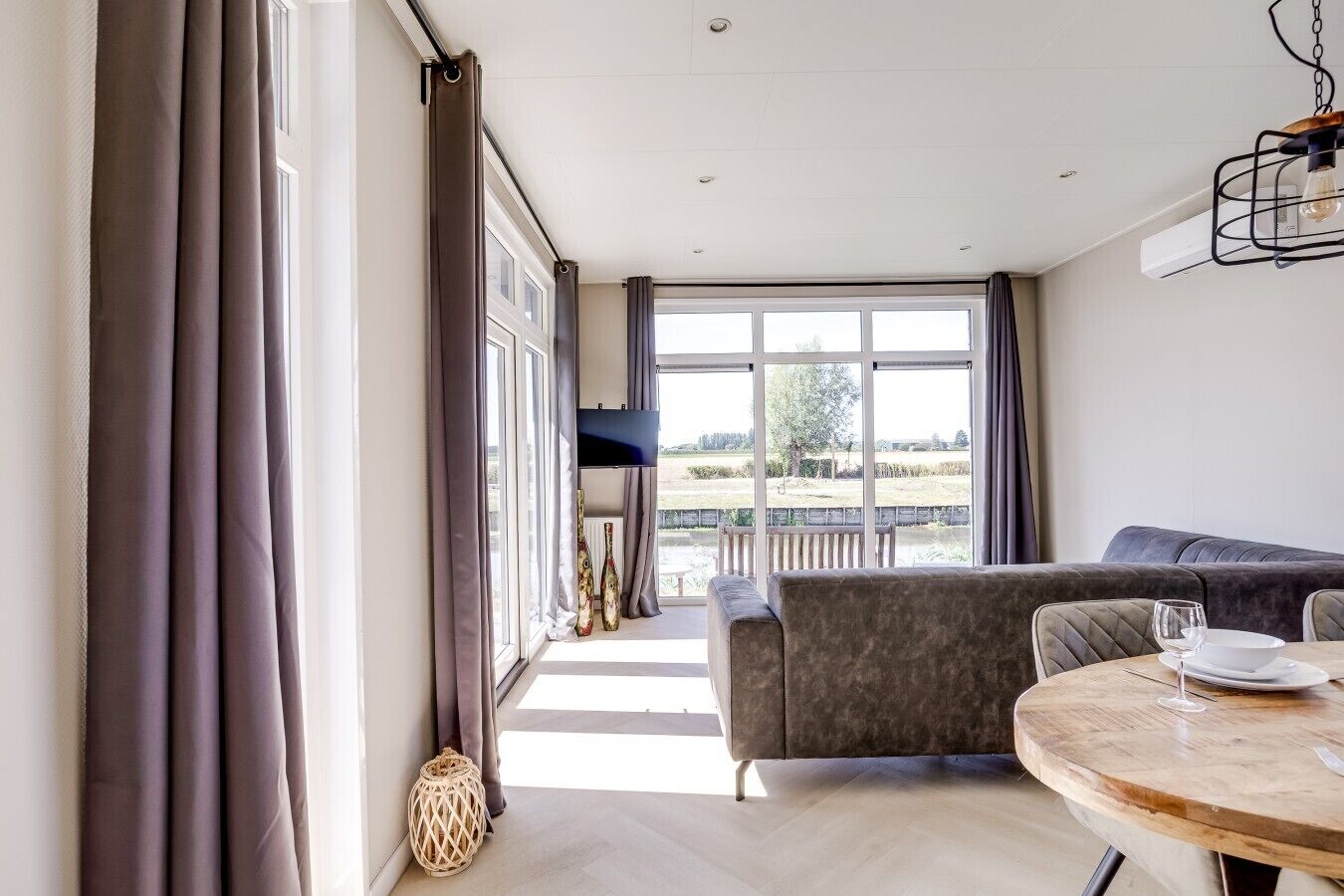 Een moderne woonkamer met een bank, een eettafel met een wijnglas, en uitzicht op een tuin door grote ramen.