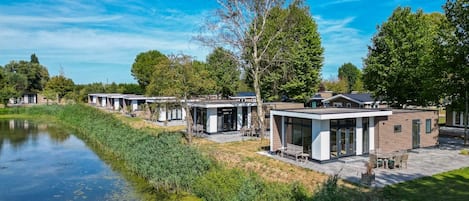 A row of modern, single-story buildings with large windows, situated beside a calm water body, surrounded by greenery.