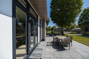 A modern outdoor patio area with a dining table and chairs, a large glass door entrance, and a well-maintained lawn. - Cube XL 71 | 4 persoons chalet in Zennewijnen (Zennewijnen)