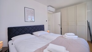 A neatly arranged bedroom with a bed, pillows, a bedside lamp, a folded towel, and a framed artwork on the wall. - City Lux Suite (Split)