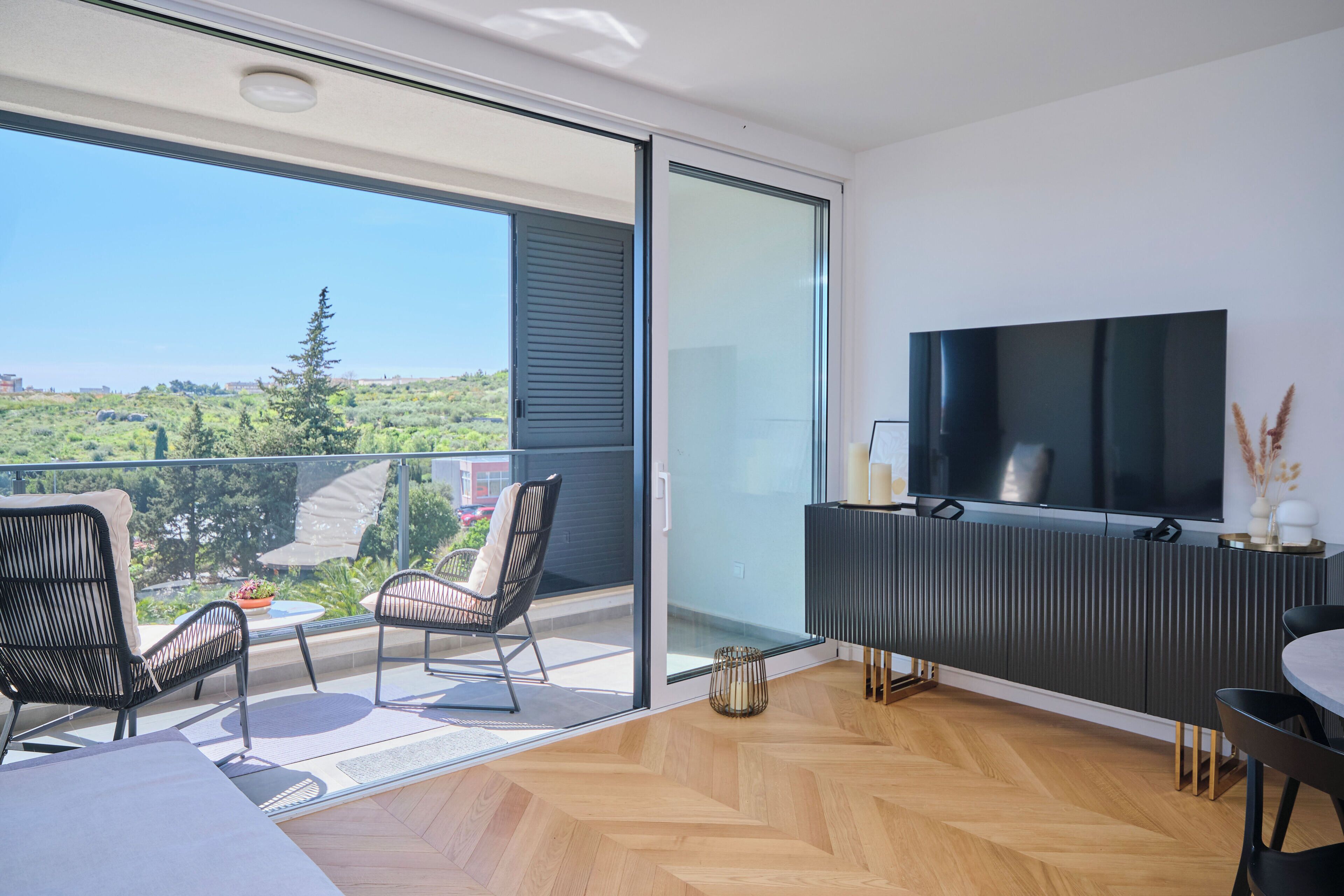 A modern balcony with a TV, seating, and a view of greenery.