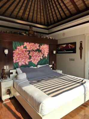 A bedroom with a bed, nightstands, a decorative headboard, a ceiling with wooden beams, and a wall with a floral painting.