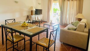 Una sala de estar con una mesa de vidrio, sillas de mimbre, un sofá, un televisor y una ventana con cortinas.