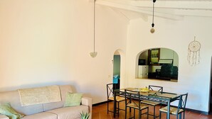 Una sala de estar con sofá, mesa de centro, mesa de comedor con sillas, una zona de cocina con refrigerador y un atrapasueños en la pared.