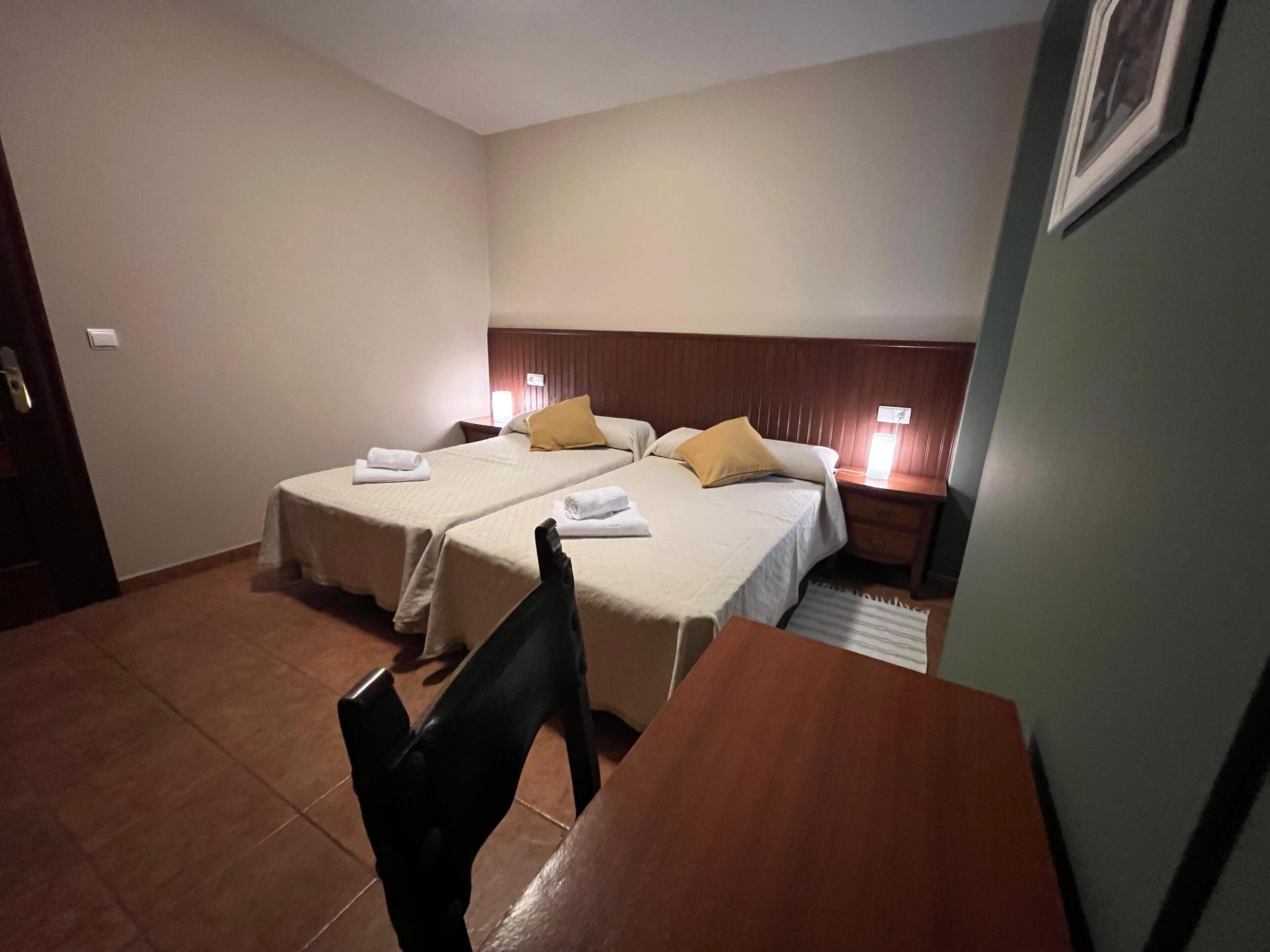 A hotel room with two beds, a wooden table, a chair, and a nightstand with a lamp.