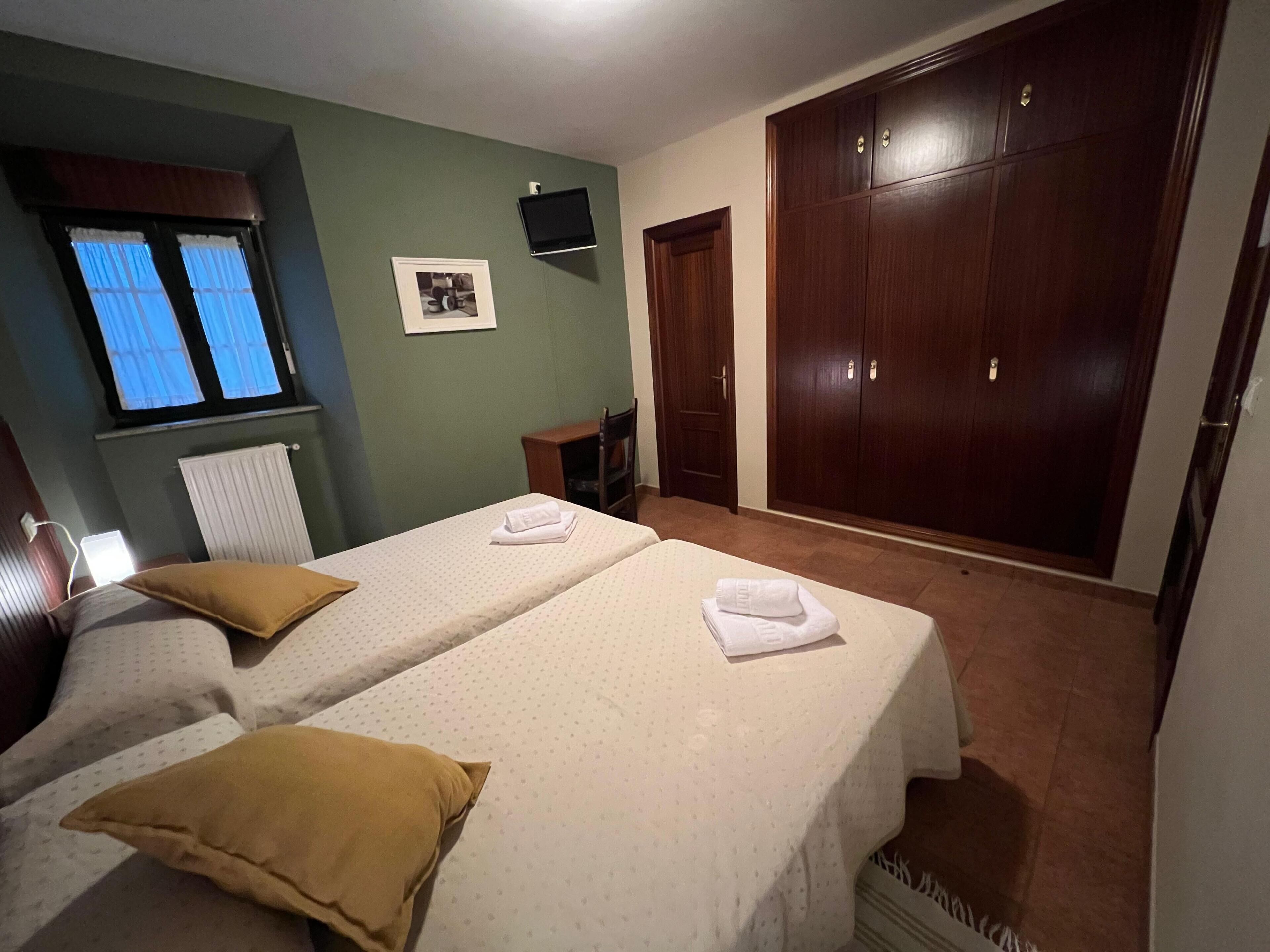 Habitación de hotel con dos camas, un armario, una mesita, una ventana con cortinas, un televisor montado en la pared y un radiador.