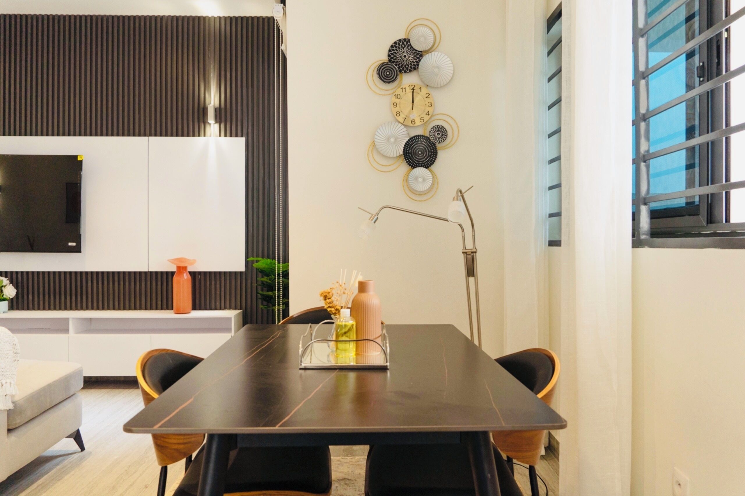 A modern dining area with a dark wooden table, two chairs, a decorative wall clock, and a mounted flat-screen TV.