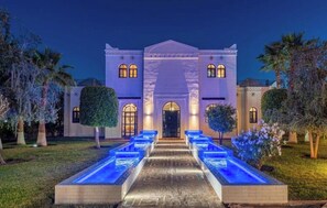 A large, well-lit building with a central entrance, surrounded by a landscaped garden and a water feature. - Palais Hadi (Tameslouht)