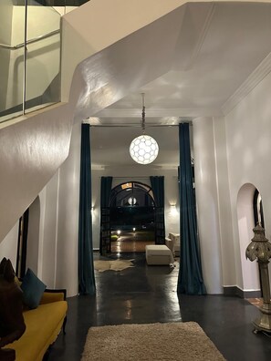 A spacious interior with a staircase, a round ceiling light, and a view into a dark room through an archway. - Palais Hadi (Tameslouht)