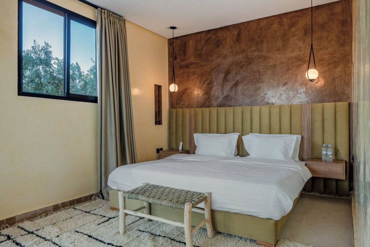 A bedroom with a bed, bedside tables, a bench, a window with curtains, and a wall with a textured finish.