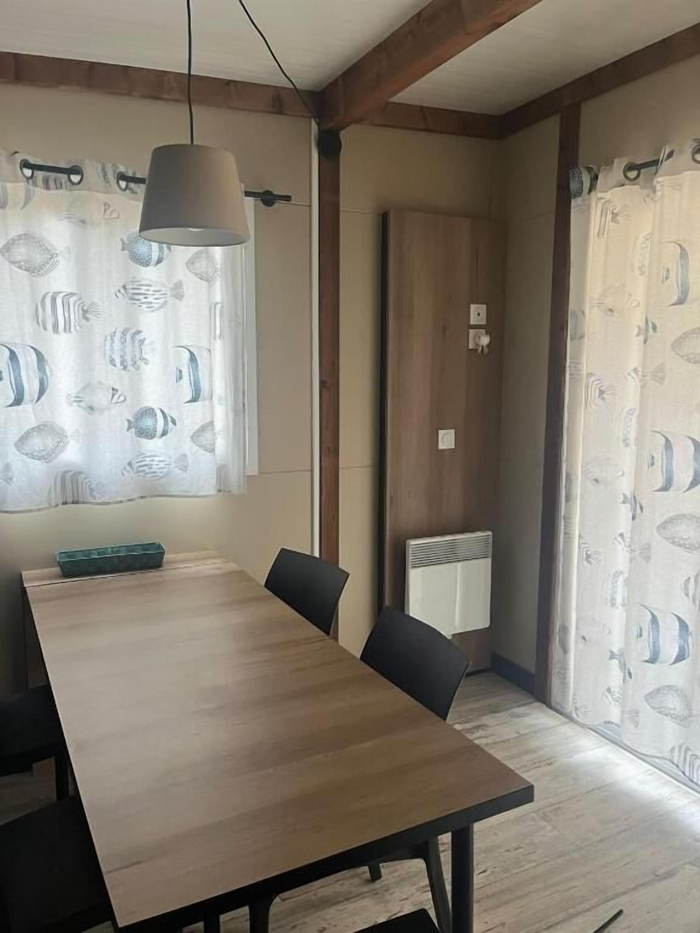 En matplats med ett träbord, svarta stolar, en hängande lampa och mönstrade gardiner.