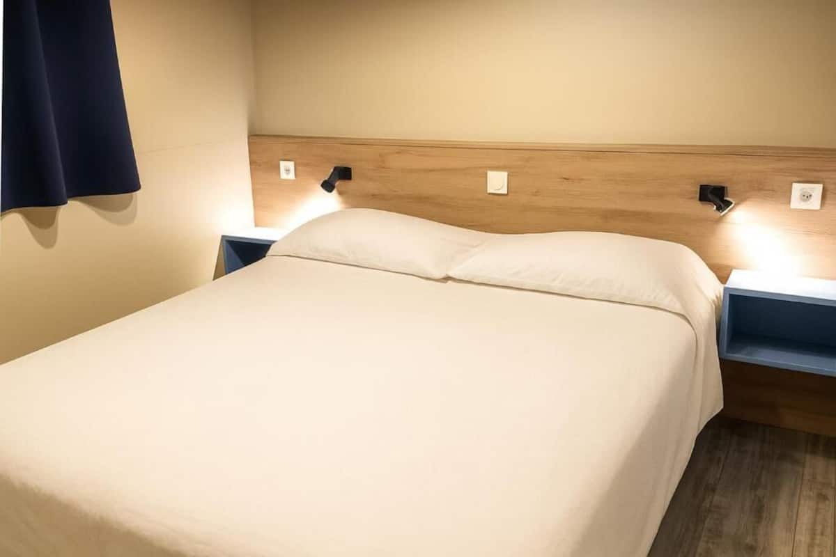 Un lit bien fait, avec une tête de lit en bois, deux tables de chevet avec des lampes et un rideau bleu foncé sur le côté gauche.