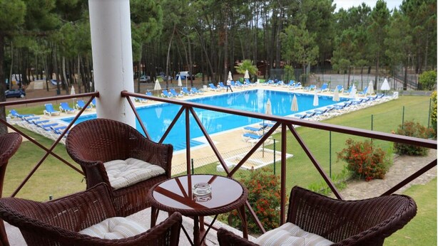 A poolside area with wicker chairs and a table, overlooking a swimming pool with lounge chairs and umbrellas.