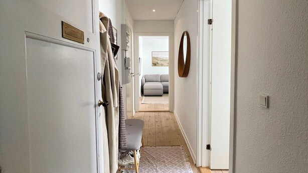 Un couloir avec un banc, un porte-manteau et une porte donnant sur une pièce où se trouve un canapé.