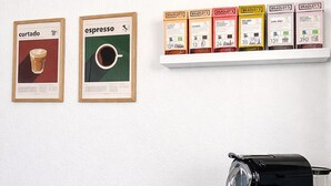 Une machine à café, deux illustrations encadrées représentant des tasses de café et une étagère contenant des capsules de café.