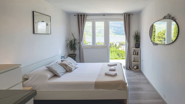 A bedroom with a bed, a bedside table, a mirror, a window with curtains, and a view of greenery outside. -  1 bedroom apt in Korčula -Cozy Island Retreat /Walk Everywhere+Private Parking/ (Korčula)