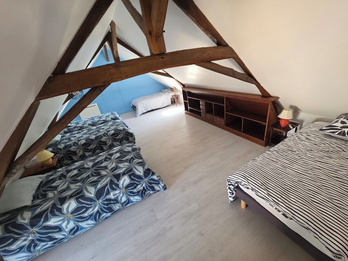 Une chambre située dans un espace de type « loft », avec un plafond en pente, des poutres en bois, un lit avec une literie à motifs, une table de chevet et un petit coin salon agrémenté d’un coussin.