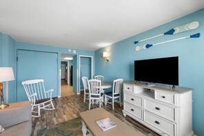 A room with a TV, white furniture, a rocking chair, and two paddleboards on the wall. - Beach and Golf 1 Bedroom Condo 9th Floor (Myrtle Beach)