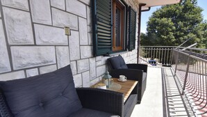 Uma varanda com móveis de vime, uma mesa com uma xícara e uma garrafa, e um muro de pedra com persianas.
