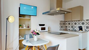 Ett litet, praktiskt kök med mikrovågsugn, brödrost och kaffemaskin, ett litet matbord med stolar och en tv som är monterad ovanför bänkskivan.