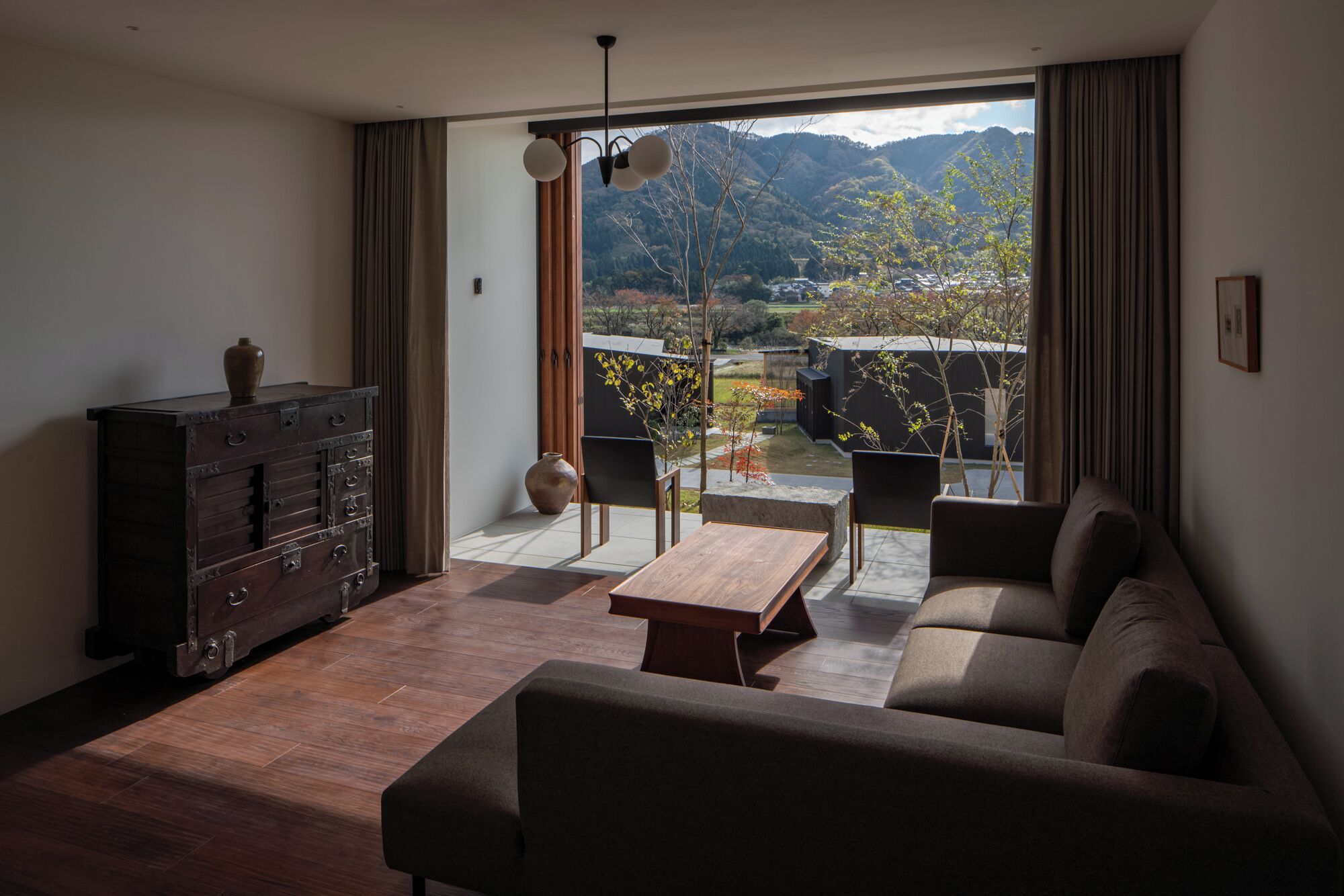A modern living room with a sofa, wooden coffee table, and a dark wooden cabinet. A large window offers a view of a mountainous landscape.