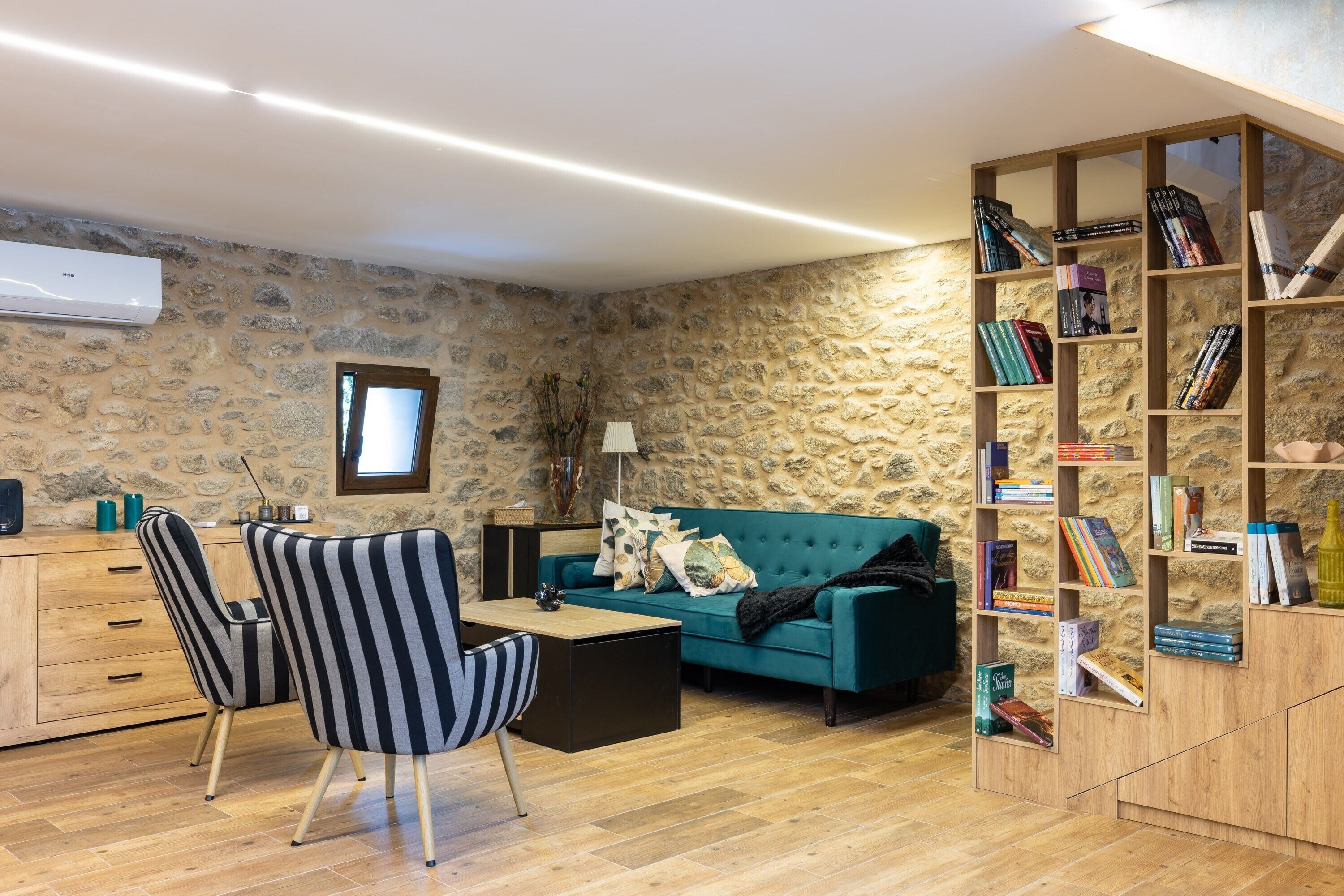 A cozy living room with a stone wall, a teal sofa, striped chairs, a wooden coffee table, and a bookshelf filled with books.